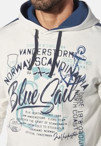 Hooded sweatshirt in cream with blue accents. Features graphic text, an anchor design, and textured inner hood. Drawstrings in dark blue.