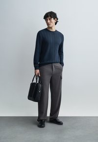 Pulover texturat bleu marin, pantaloni largi gri cu buzunare și geantă tote neagră. Pantofii negri completează ținuta. Design simplu și modern.