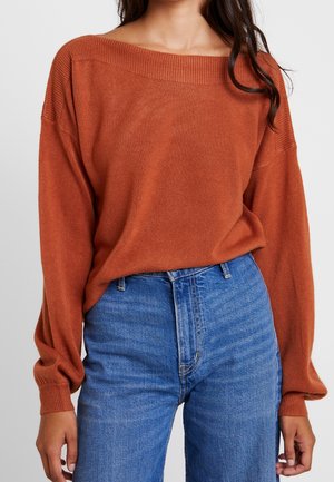 Jersey naranja óxido con hombros descubiertos y mangas largas abullonadas combinado con vaqueros azules de cintura alta en una mujer.