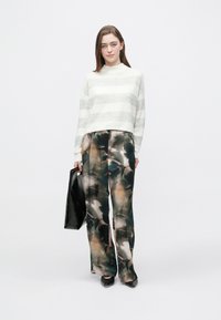 Pulover gri deschis cu dungi albe și guler înalt, asortat cu pantaloni largi, cu model multicolor. Modelul ține o geantă neagră.