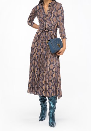Donna che indossa un lungo vestito con stampa serpente, tiene in mano una borsa trapuntata blu e indossa stivali lucidi color turchese fino al ginocchio.