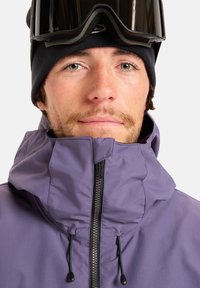 Uomo con berretto nero e occhiali da sci sulla fronte, giacca viola zipata con coulisse nera, espressione neutra.