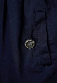 Laivastonsinistä kangasta, jolla on sileä pinta, varustettuna nelireikäisellä kirkkaalla napilla, joka sijaitsee sivutaskun yläpuolella.