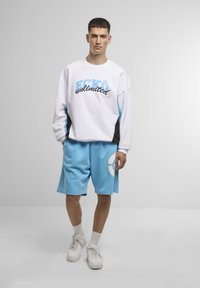 Felpa bianca con accenti blu e neri, con testo "ECKO unlimited". Pantaloni corti azzurri con una grafica a cerchio bianco. Sneakers bianche.