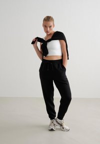 Schwarze Jogginghosen mit elastischem Bund und Taschen, kombiniert mit einem weißen, kurzen Tanktop und einem schwarzen Pullover, der über eine Schulter gelegt ist.