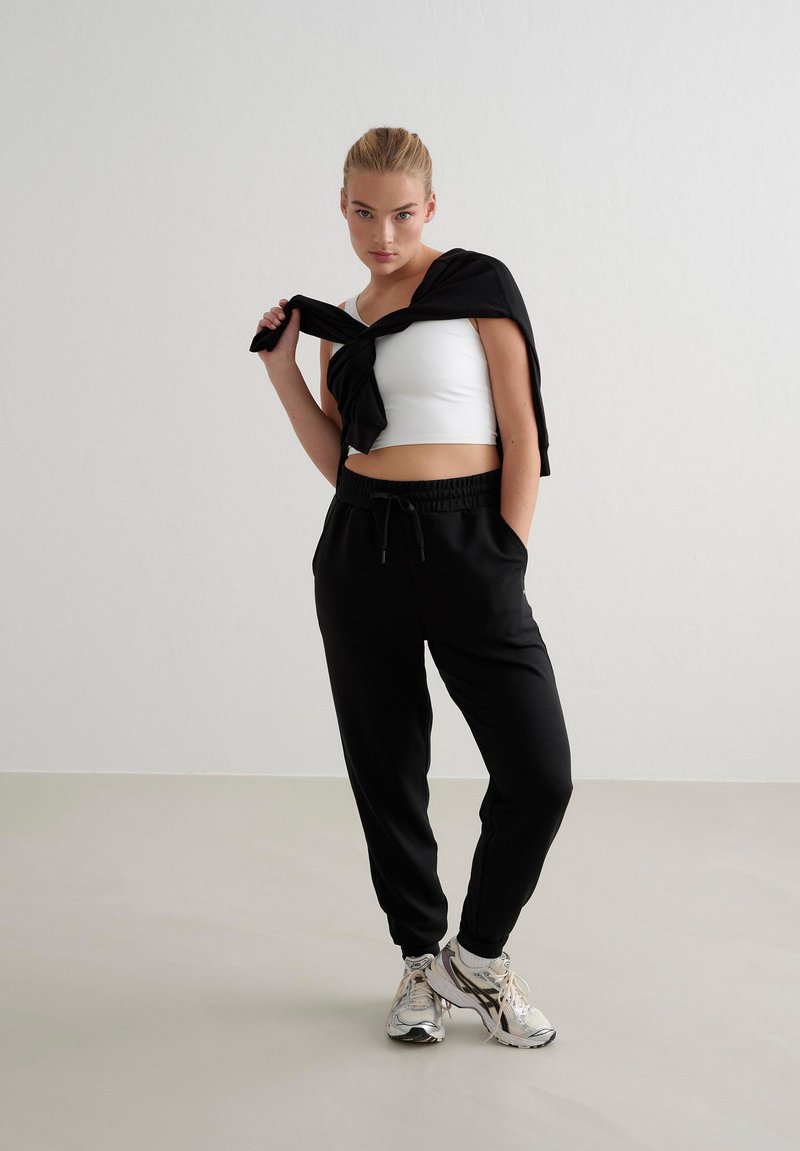 Schwarze Jogginghosen mit elastischem Bund und Taschen, kombiniert mit einem weißen, kurzen Tanktop und einem schwarzen Pullover, der über eine Schulter gelegt ist.