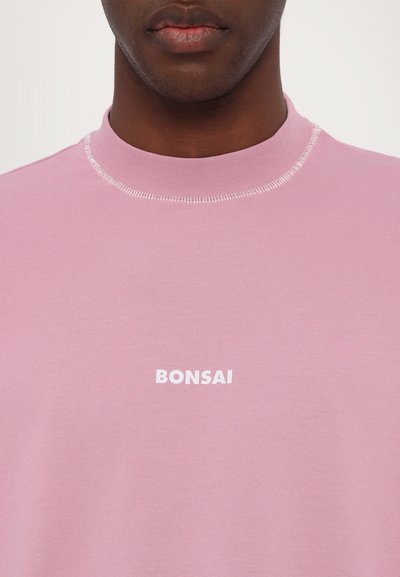 Gaiši rozā sporta krekls ar augstu apkakli, īsām piedurknēm un baltu "BONSAI" logotipu centrā uz krūtīm. Gluda auduma tekstūra.