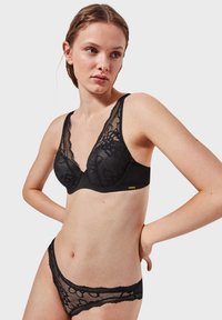 Reggiseno in pizzo nero con motivo floreale e coppe trasparenti, caratterizzato da un profondo scollo a V e spalline regolabili. Slip in rete nera con rifiniture in pizzo.