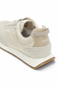 Zapatilla de cuero beige con parte superior lisa, puntera redondeada, costuras tonales y suela de goma. Cuenta con detalle de logo y una lengüeta en el talón.