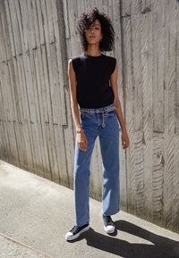 Top noir sans manches, jean bleu à jambes larges avec ceinture chaîne, et baskets montantes noires. En arrière-plan, un mur en bois texturé.