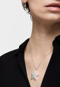 Ciondolo a forma di stella argentata su una collana a catena, con una superficie liscia e riflettente, indossato sopra una blusa nera con colletto.