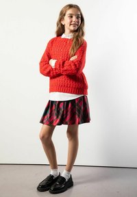 Czerwony dziergany sweter na białej koszuli z kołnierzykiem, połączony z czarno-czerwoną plisowaną spódnicą w kratę. Czarne mokasyny i białe skarpetki dopełniają wygląd.