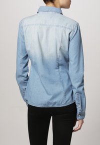Chemise en denim bleu clair avec col, poignets à boutons, et dos légèrement délavé. Présente une coupe ajustée et une texture lisse.