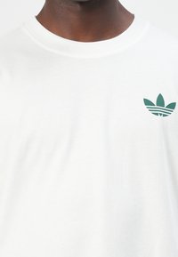 Tricou alb din bumbac cu guler rotund texturat. Prezintă un logo stilizat verde pe partea stângă a pieptului, creând un design minimalist.