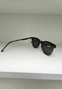 Zwarte zonnebril met een schildpadpatroon op de pootjes, ronde lenzen en dunne steunen. Gemarkeerd met "KOMONO" en CE-keurmerk.