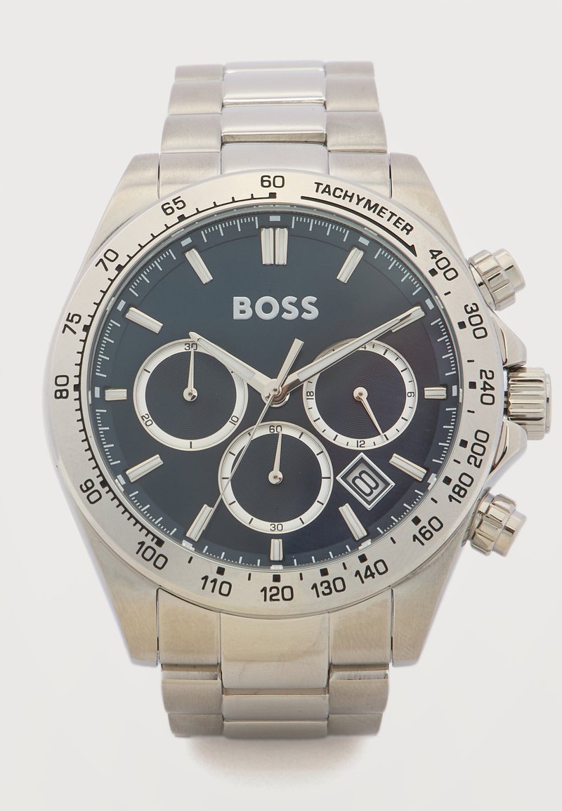 Ασημένιο μεταλλικό ρολόι χειρός με μαύρο καντράν, τρεις υποκαντράν, στεφάνη ταχυμέτρου, παράθυρο ημερομηνίας στη θέση 4 η ώρα και λογότυπο BOSS στη θέση 12 η ώρα.