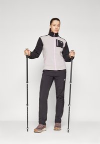 Hallide ja must fleece-jacket roosade aktsentidega, lukustatavad taskud; koos mustade pükste ja matkäbootsidega, trekke kepid käes.
