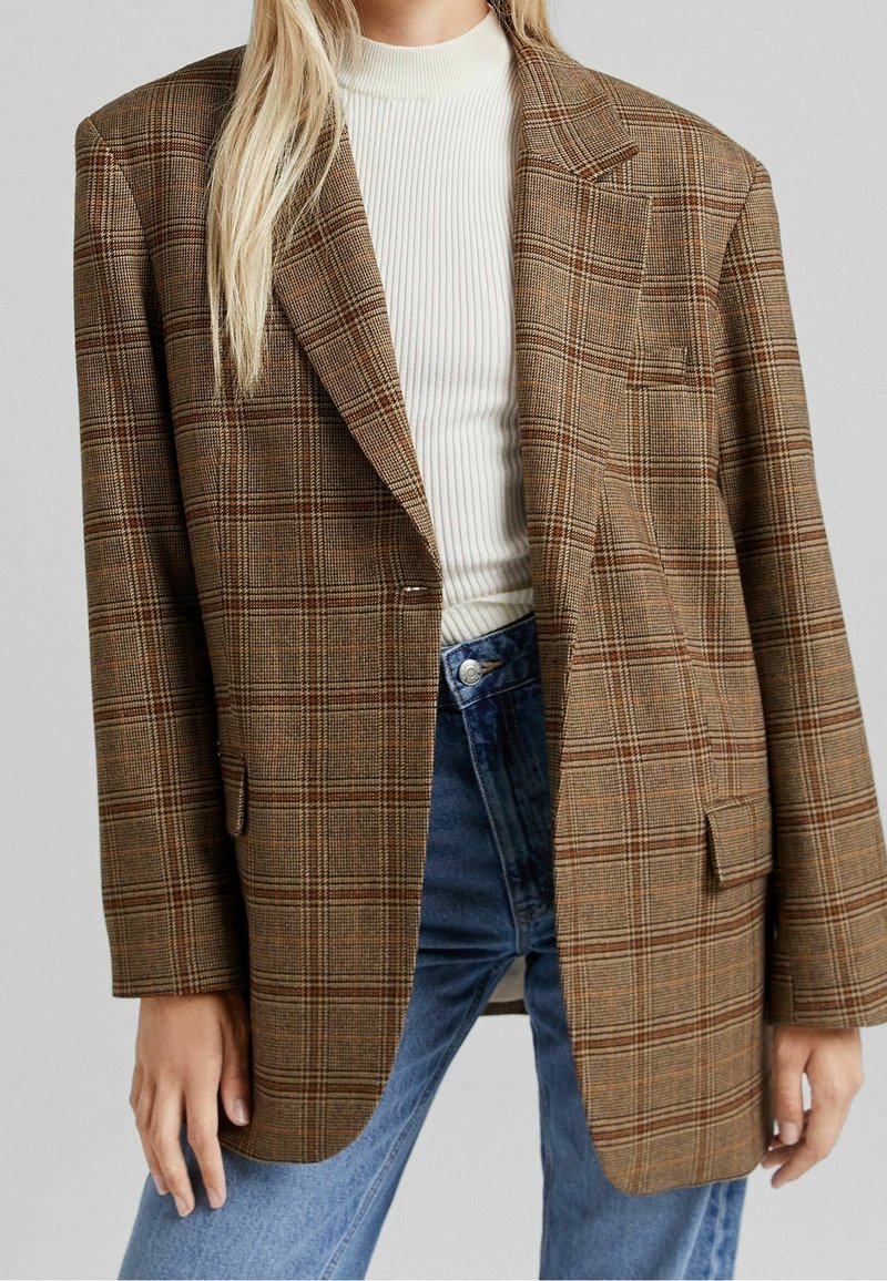 Femme portant un blazer marron à carreaux sur un col roulé blanc côtelé et un jean bleu taille haute, debout devant un fond uni.