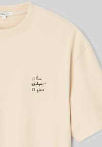 T-shirt beige avec col ras du cou, manches courtes, et design imprimé en noir représentant une liste de contrôle avec un mot barré. Matière en coton doux.