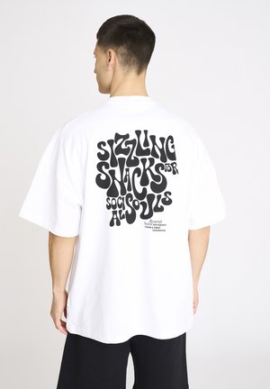 Mand iført hvid oversized t-shirt med stor sort forvrænget tekst på ryggen, der lyder "Sizzling snacks for social souls."