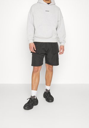 Personne portant un sweat à capuche gris clair avec une poche avant, un short cargo noir, des chaussettes blanches et des baskets noires, debout sur fond blanc.