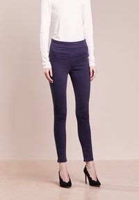 Lila smala byxor med hög midja, slät textur och detaljerad sömnad, kombinerat med svarta högklackade skor och en vit långärmad topp.