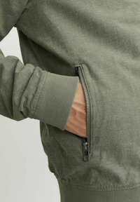 Grüne Zip-Jacke aus strukturiertem Stoff, mit gerippten Bündchen und Seitentasche mit silbernem Beschlag. Nahaufnahme des Ärmels und des Reißverschlusses.