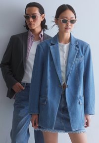 Hele sinine denim bleiser, millel on ees taskud, koos valge pluusi ja frantsiga denim seelikuga. Tumedat ülikonna jakk on taustal.