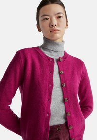 Fuchsiový pletený cardigan s kulatým výstřihem a pěti hnědými knoflíky, navlečený přes světle šedý rolák. Měkká textura s žebrovanými manžetami.