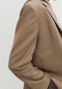 Nærbillede af en tan uldfrakkeærme og skulder, med hånden stukket ned i lommen, mod en ensfarvet lys baggrund.