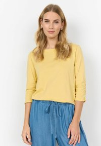 Langsleeve top in zachtgeel katoen, met een vierkante halslijn en geraffineerde details op de mouwen, gecombineerd met lichtblauwe broek met touwtjes.