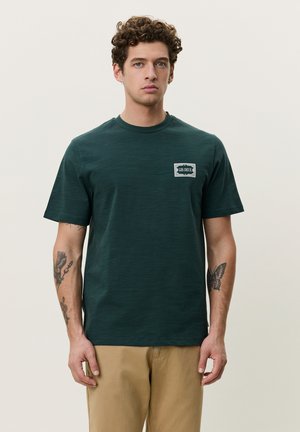 Jeune homme aux cheveux bouclés portant un t-shirt vert foncé "LES DEUX" et un pantalon beige, montrant des tatouages sur les deux avant-bras, sur un fond uni.