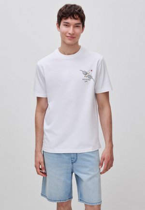 Junger Mann mit kurzen dunklen Haaren, der ein weißes T-Shirt mit einem Kassettenspieler-Motiv und hellblaue Jeansshorts trägt, steht und lächelt.