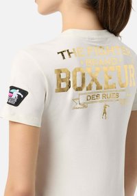 Weißes T-Shirt mit kurzen Ärmeln und strukturiertem Gold- und Schwarzdruck auf der Rückseite, mit dem Text "THE FIGHTER BRAND BOXEUR DES RUES" und einem Aufnäher am Ärmel.