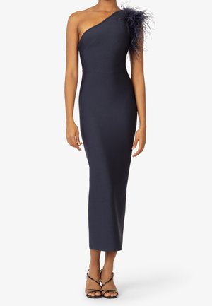 Mujer con un vestido ajustado azul marino con un solo hombro, detalle de plumas en el hombro y tacones negros de tiras.