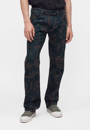 Homme portant un jean noir et marron usé avec des baskets en toile verte et une chemise noire sur fond blanc.