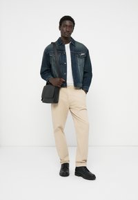 Jaqueta de denim com um acabamento azul desbotado, t-shirt branca, calças beiges e botas pretas de atacador, segurando uma mala messenger preta.