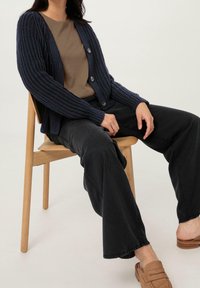Cardigan en velours côtelé bleu marine avec de gros boutons, porté sur une chemise marron. Pantalons larges noirs et chaussures à enfiler beiges. Assis sur une chaise en bois.