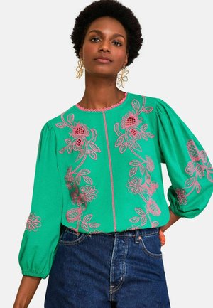 Vrouw draagt een felgroene blouse met roze bloemrijke borduursels en opgeblazen mouwen, gecombineerd met blauwe jeans met hoge taille.