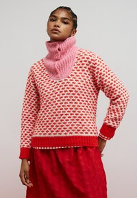 Roter und weiß gemusterter Pullover mit einem rosa gerippten Kragen und Knöpfen. Der untere Saum und die Ärmelbündchen sind einfarbig rot.