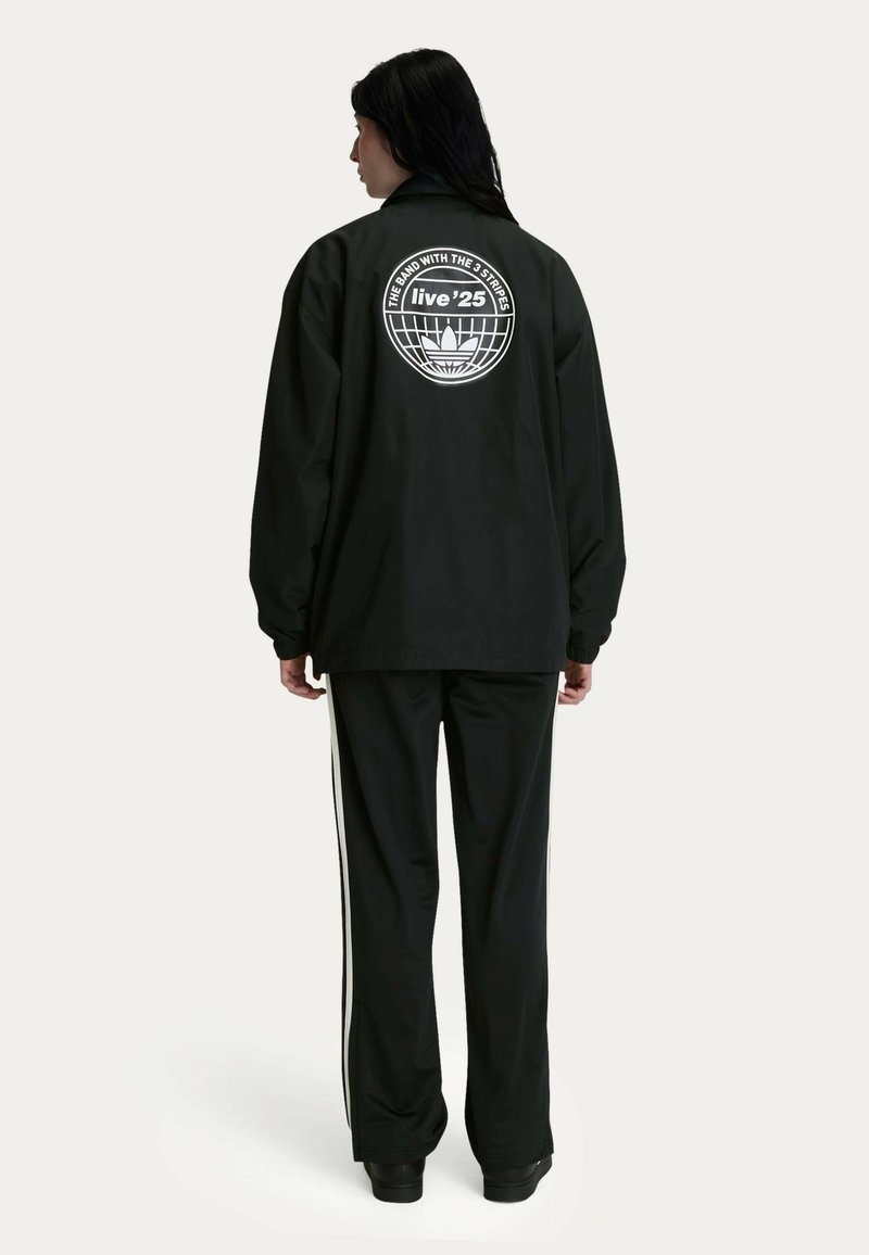 Black windbreaker with a large white graphic on the back, paired with black track pants featuring white side stripes. Standard fit, smooth texture.