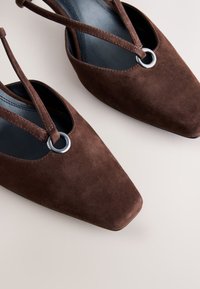 Chaussures slingback en daim marron à bout pointu avec détail anneau argenté et doublure intérieure noire, présentées sur une surface claire.