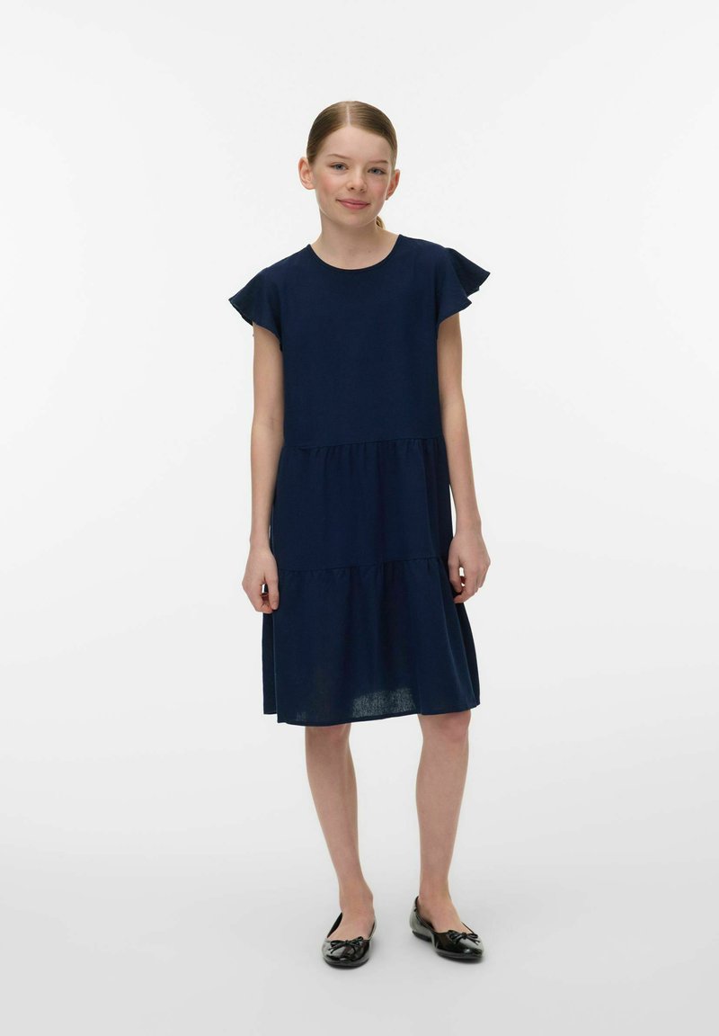 Meisje staat met ontspannen handen, draagt een marineblauwe gelaagde jurk en zwarte ballerina's, tegen een effen witte achtergrond.