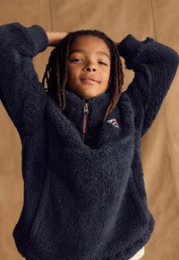 Pull-over en polaire marine avec une demi-fermeture éclair, un col haut et des poignets côtelés. Présente un écusson logo et un matériau texturé et doux.