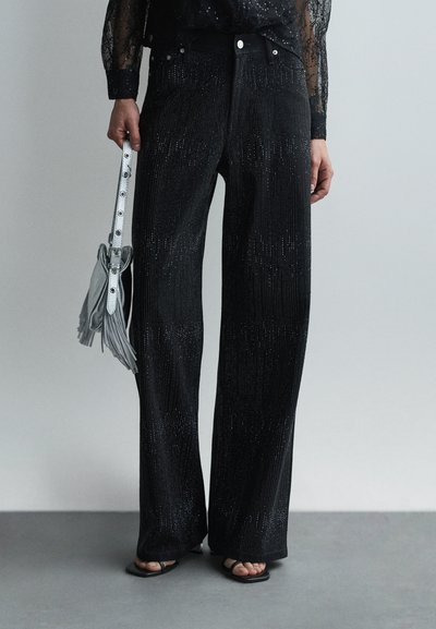 Zwarte, wijde broek met een metallic glans, voorzien van verticale strepen en een hoge taille, gecombineerd met een lichtgrijze fringe handtas.