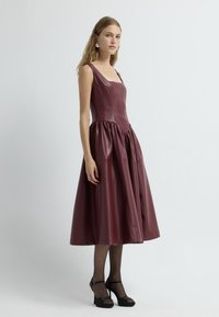 Vestido de cuero burdeos con un corpiño ajustado y falda acampanada, que presenta un escote cuadrado y detalles plisados, combinado con tacones negros.