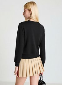 Sweat-shirt noir à manches longues, col rond et ourlet côtelé, associé à une jupe plissée beige. Le modèle tient un sac à main noir.