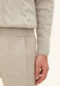 Light beige cable-knit sweater with ribbed hem and cuffs, paired with light beige textured trousers, displaying a soft woolen material.