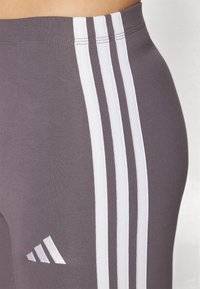Leggings atléticos cinzentos com um design de três riscas brancas ao longo do lado e um pequeno logotipo branco. O tecido parece liso e ajustado.