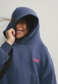 Sudadera azul marino con una gran capucha, que cuenta con un bolsillo canguro en la parte delantera y un logo "SuperDry" rosa en el pecho. Tejido suave y texturizado.